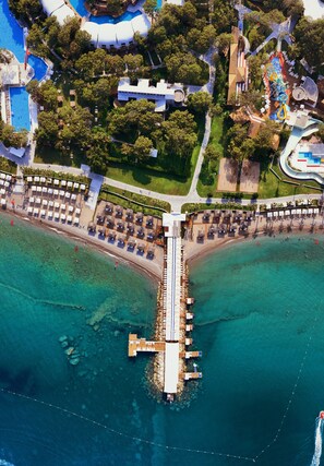 An aerial view of a coastal resort with a pier extending into turquoise waters, surrounded by lush greenery and a variety of amenities including a pool, beach area, and shaded seating.