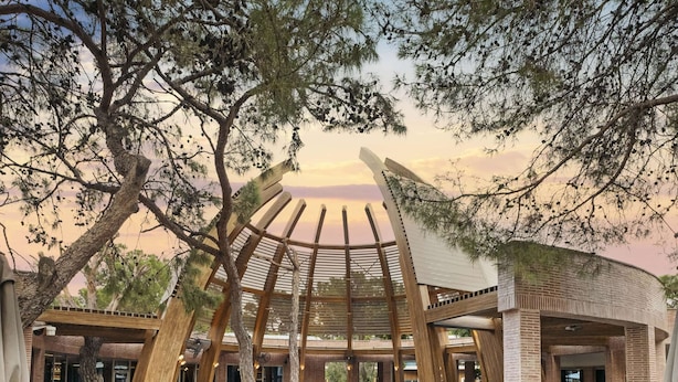 Zona de menjador a l'aire lliure amb taules i cadires de fusta, envoltada d'arbres i una piscina.
