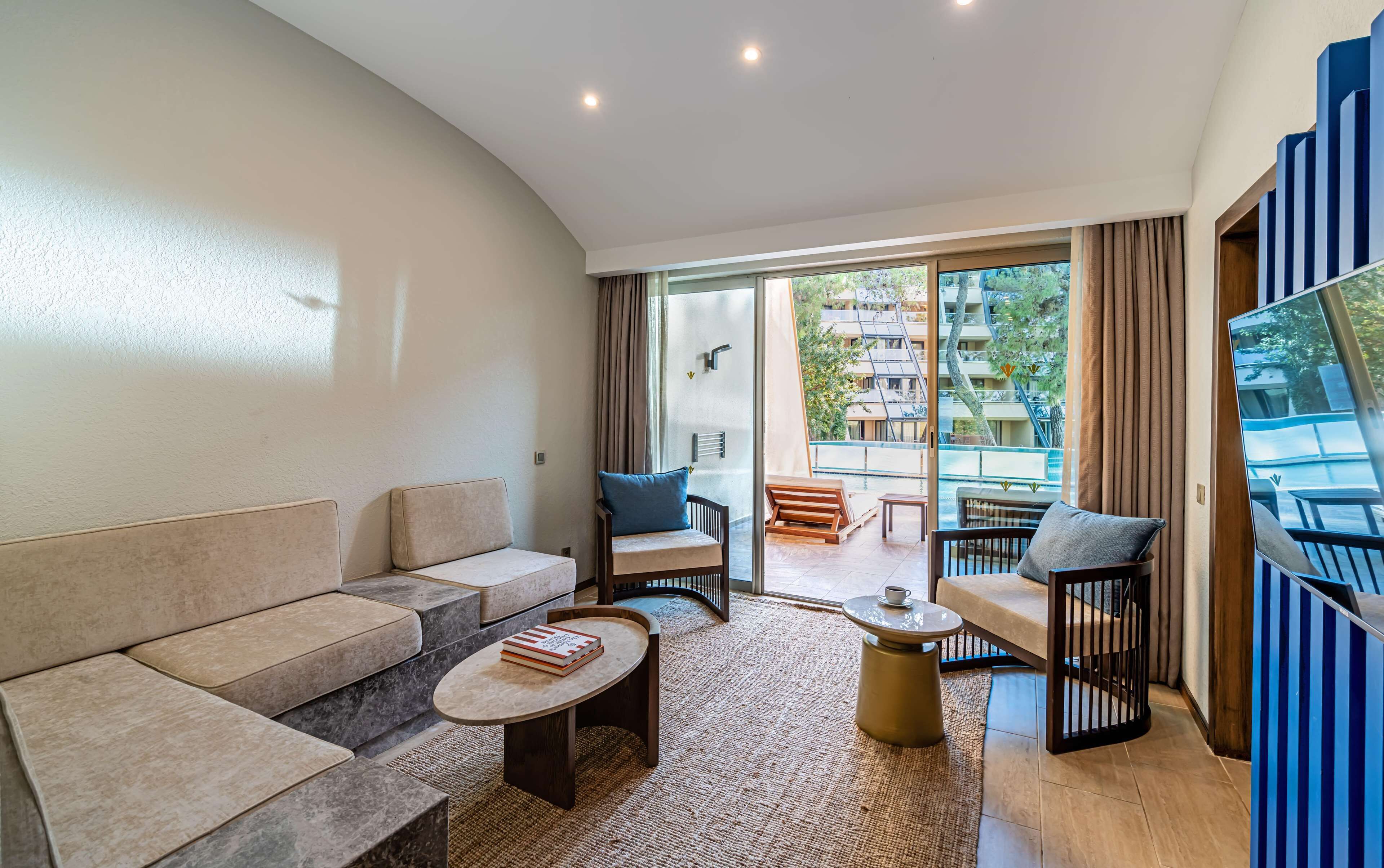A modern living room with a sofa, armchairs, a coffee table, and a view of a pool and buildings outside.