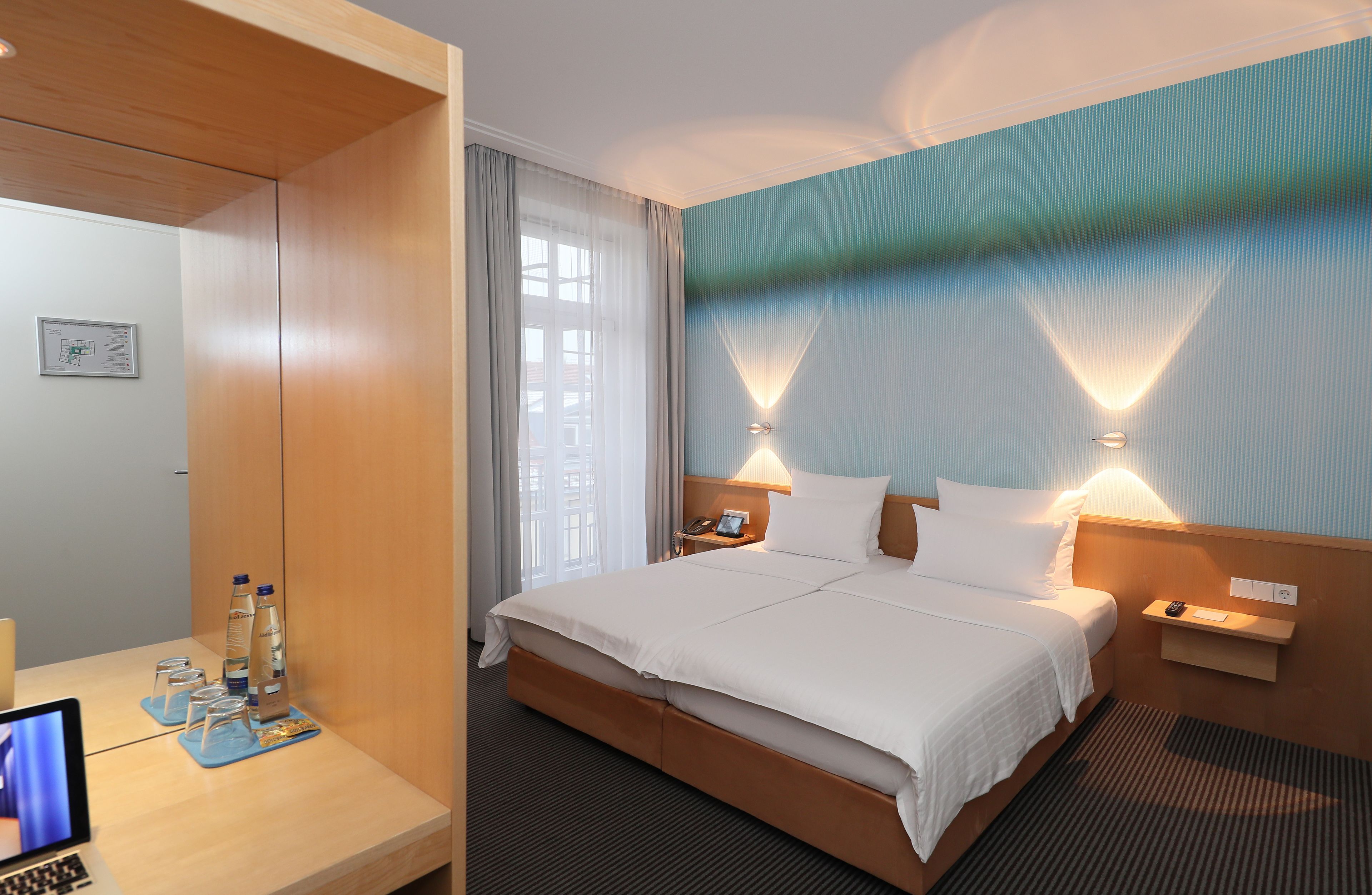 A hotel room with a large bed, bedside tables, a desk with a laptop, and a view of the city through the window.