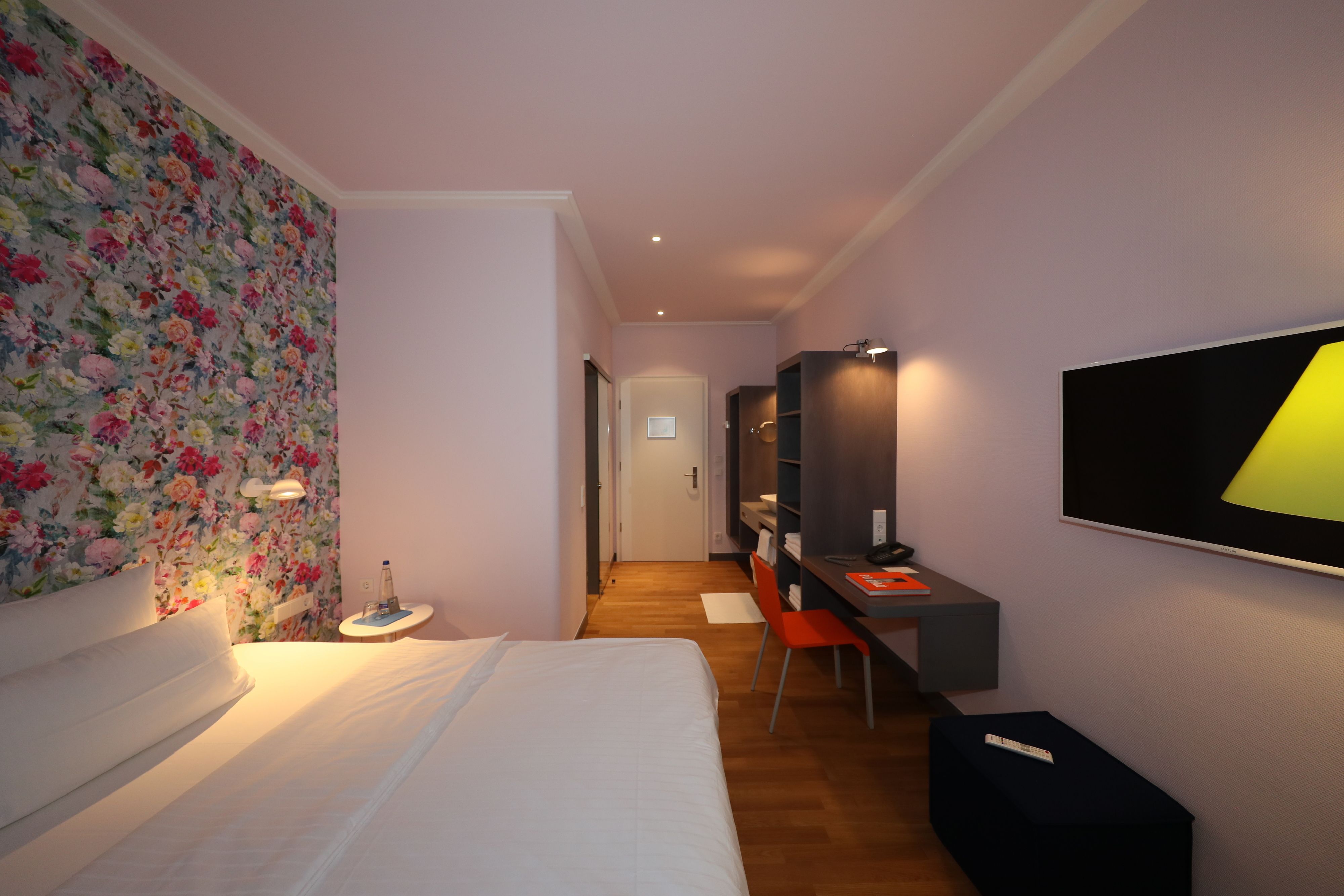 Habitació d'hotel amb un llit, un escriptori amb una cadira, un mirall, una televisió i una paret amb paper pintat floral.