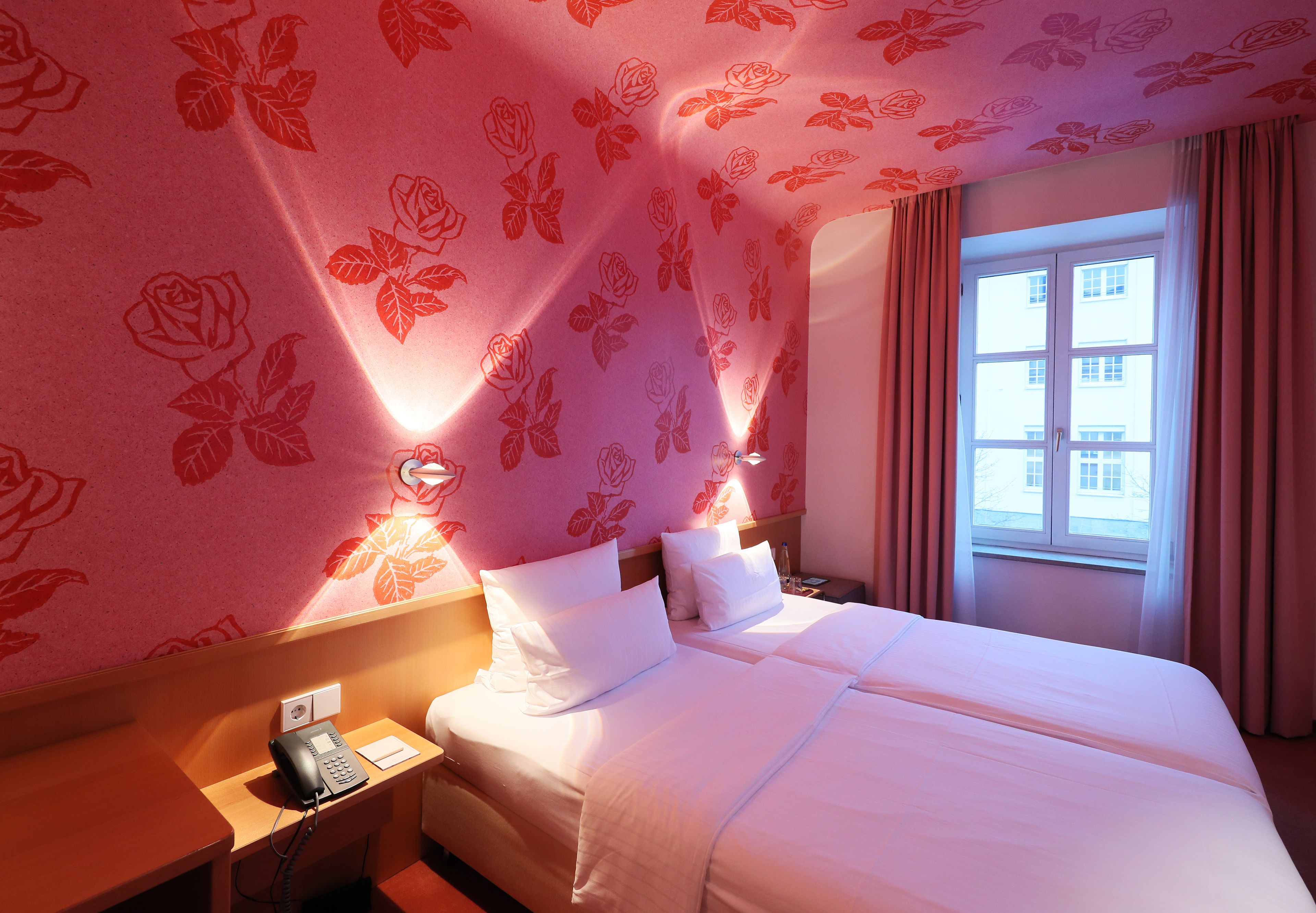 Quarto de hotel com papel de parede floral, uma cama com roupa de cama branca, mesa de cabeceira com telefone e janela com cortinas.