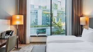 Quarto de hotel com uma cama grande, duas luminárias de cabeceira, uma mesa com cafeteira e vista para um edifício e área verde lá fora.