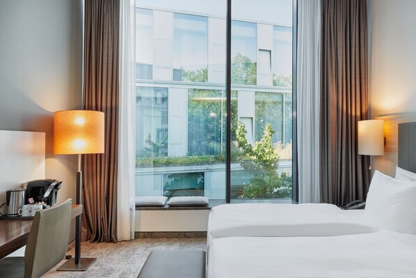Habitación de hotel con una cama grande, dos lámparas de noche, un escritorio con cafetera y vistas a un edificio y zonas verdes.