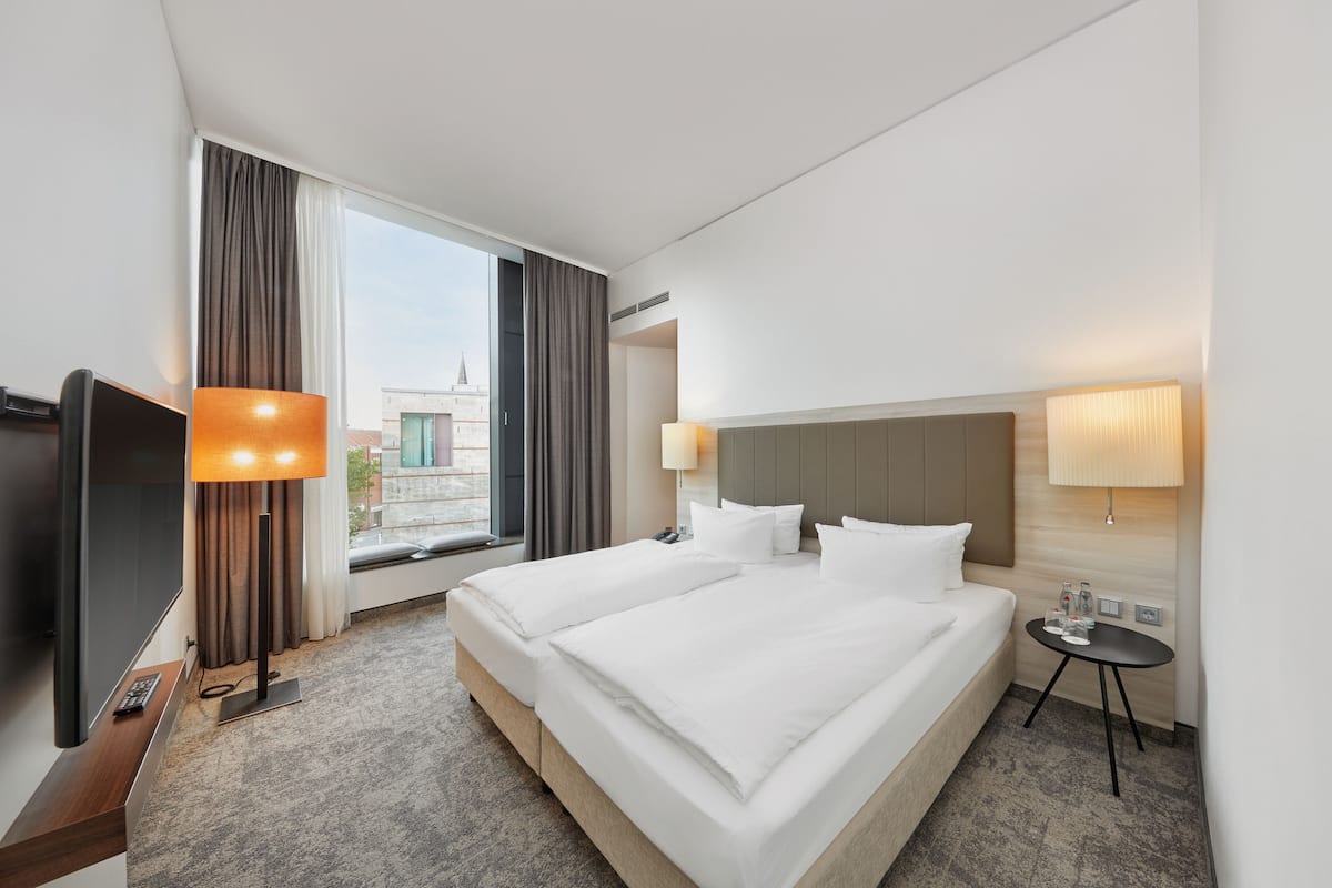 A hotel room with a large bed, two bedside tables, a TV, and a window with curtains.