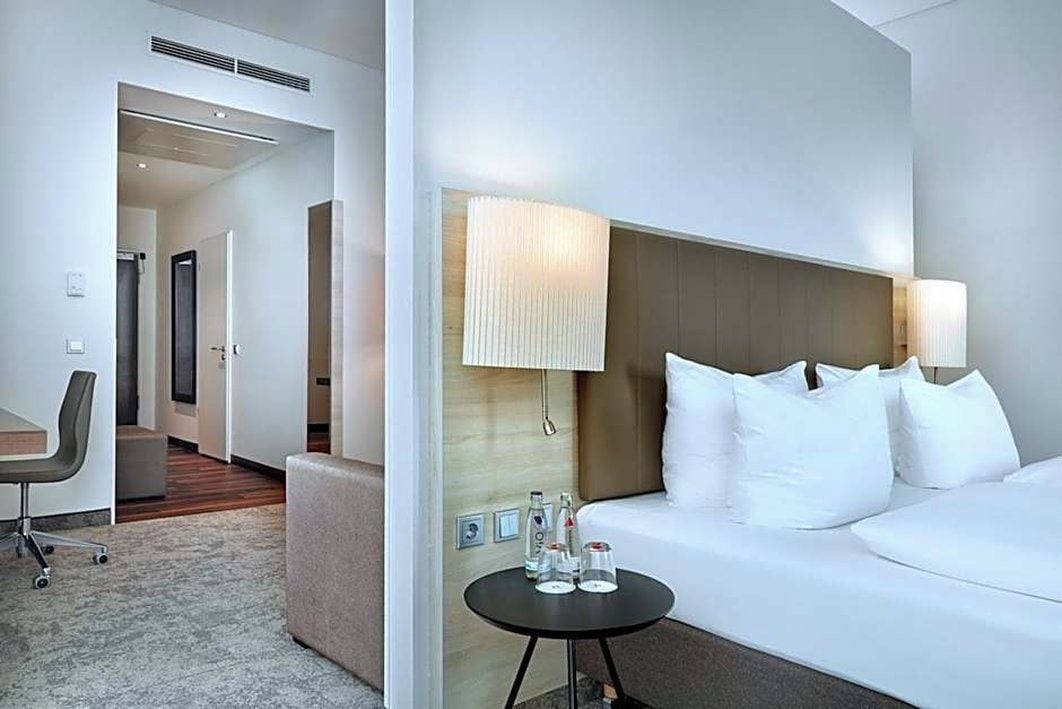 A hotel room with a bed, bedside lamps, a small table with water bottles, a chair, and a desk.