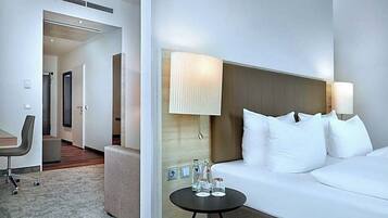 A hotel room with a bed, bedside lamps, a small table with water bottles, a chair, and a desk.