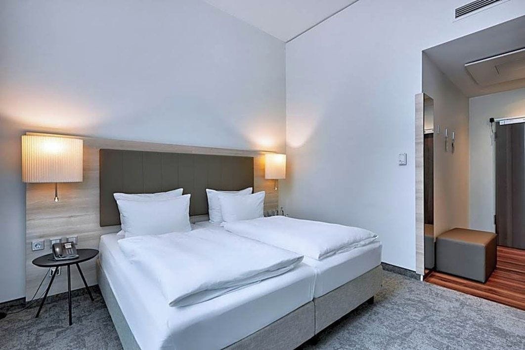 A hotel room with a double bed, two bedside lamps, a small table, and a bench.
