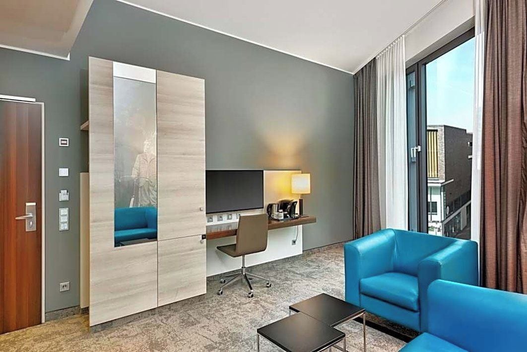 Quarto de hotel com uma janela grande, uma escrivaninha com cadeira, um sofá e uma máquina de café.