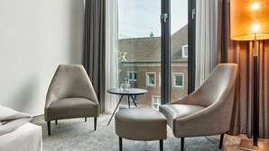 Habitación de hotel con dos sillones, una mesita redonda, una lámpara y vistas a un edificio de ladrillo a través de amplias ventanas.