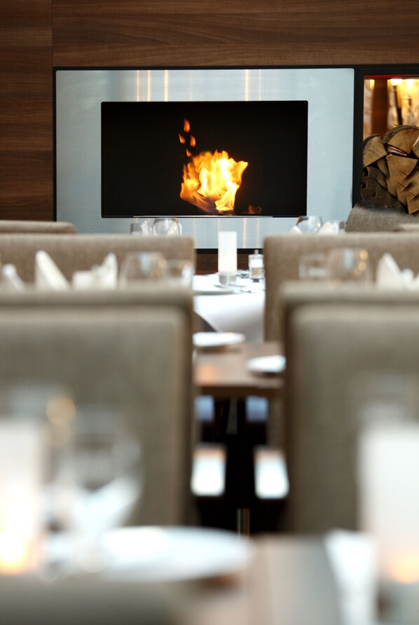 A modern dining area with a television displaying a fireplace, comfortable seating, and a neatly arranged table with glasses and cutlery. - Hotel Münster-Altstadt (Münster)