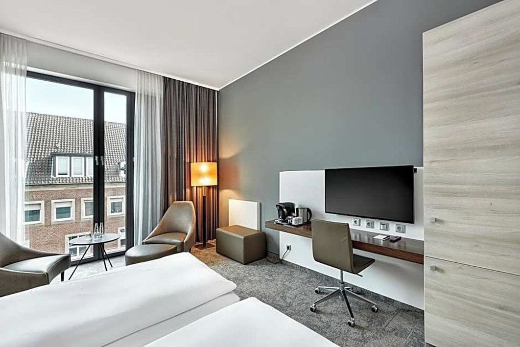 Ein Hotelzimmer mit großem Fenster, einer Sitzecke, einem Schreibtisch mit Flachbildfernseher, einer Lampe und einer Kaffeemaschine.