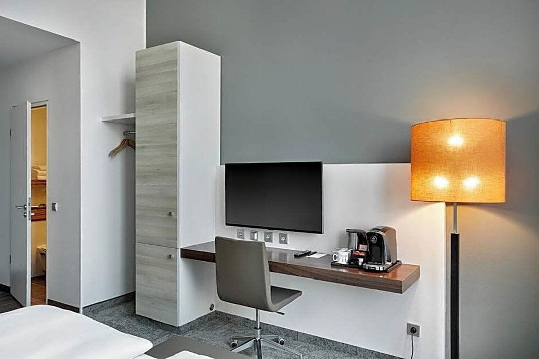Ett hotellrum med en säng, ett skrivbord med en platt-TV, en lampa och en kaffebryggare.
