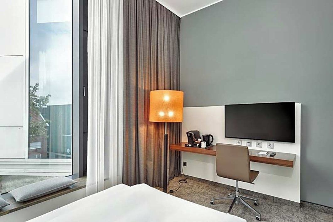 A modern hotel room with a large window, a flat-screen TV, a desk with a lamp, and a comfortable bed.