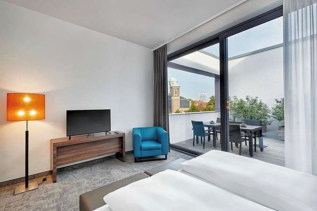 A hotel room with a bed, a TV, a desk, a chair, and a balcony with a view of a building.