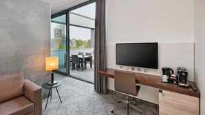 Habitación de hotel con escritorio, silla, televisor, lámpara, cafetera y vistas al exterior.