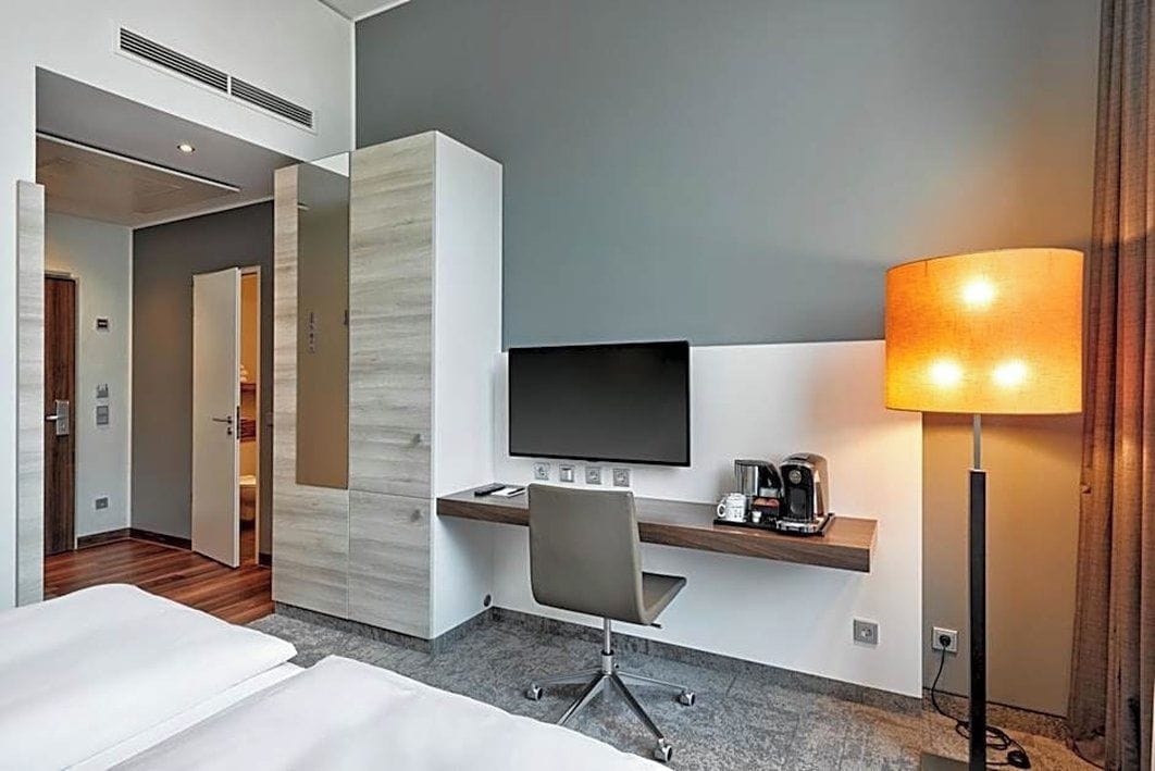 Habitación de hotel con cama, escritorio con silla, televisión de pantalla plana, lámpara y cafetera.