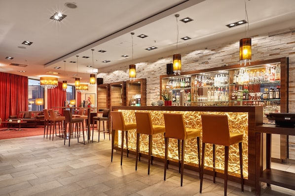 A bar area with a wooden counter, multiple bar stools, and overhead lighting. - Hotel Münster-Altstadt (Münster)