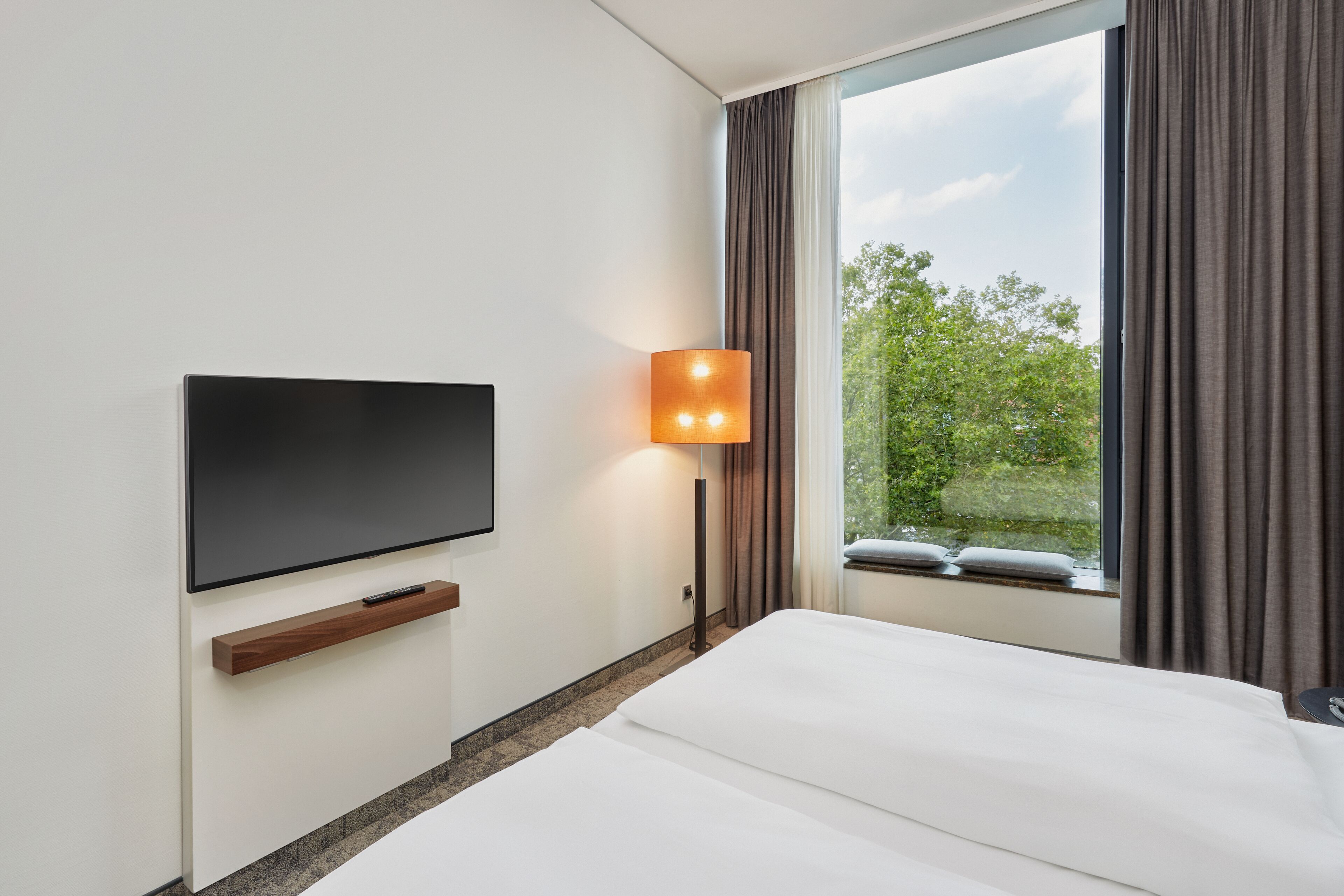 A hotel room with a flat-screen TV mounted on the wall, a bedside table with a remote control, a lamp, and a large window with curtains.