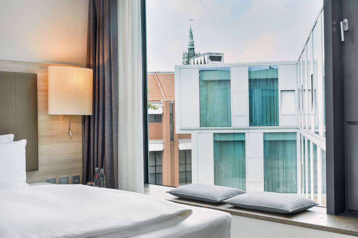 A hotel room with a large bed, a bedside lamp, a view of a modern building, and a window with curtains.