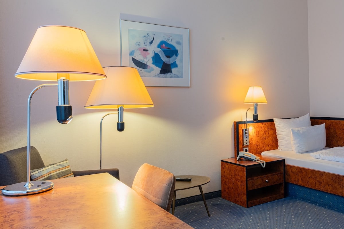 A hotel room with a bed, two bedside lamps, a chair, a small table, a nightstand with a phone, and a framed artwork on the wall.