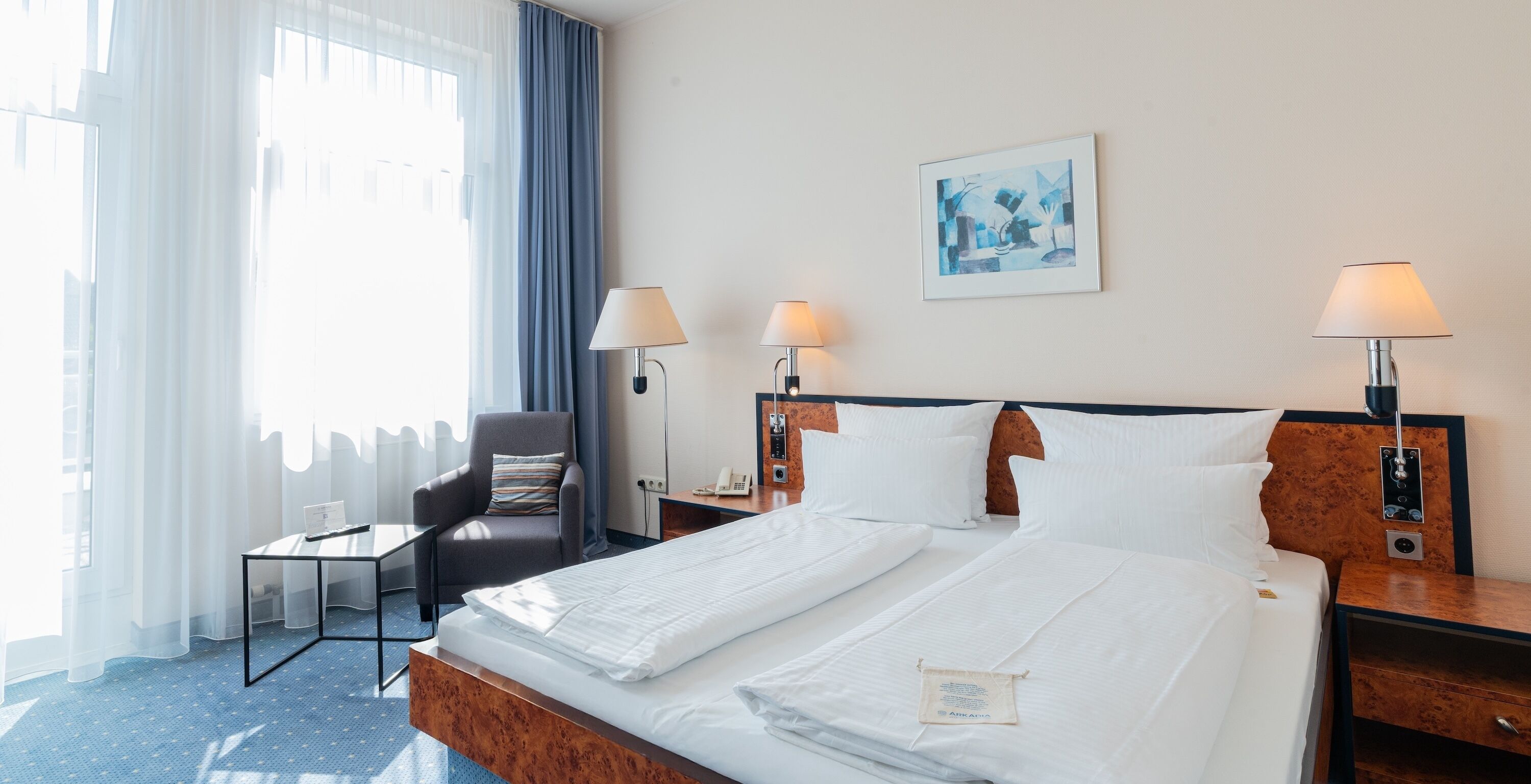 A hotel room with a bed, two bedside tables with lamps, a chair, a desk, and a window with curtains.