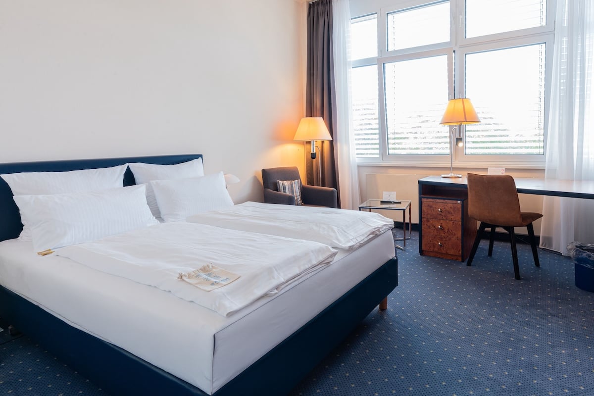 A hotel room with a large bed, two bedside lamps, a desk, a chair, and a window with blinds.