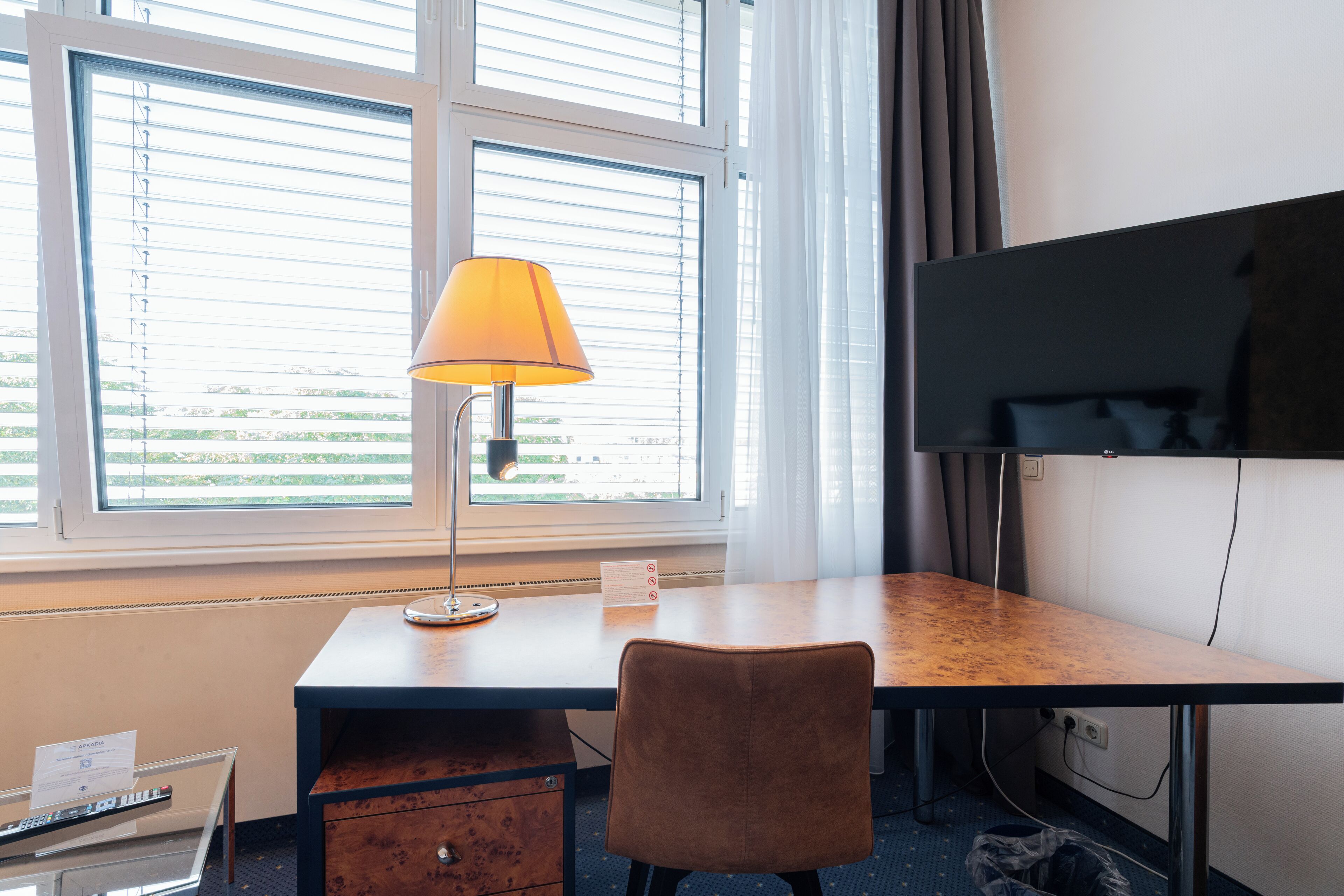 Habitación de hotel con escritorio, silla, lámpara, televisión y ventana con persianas.