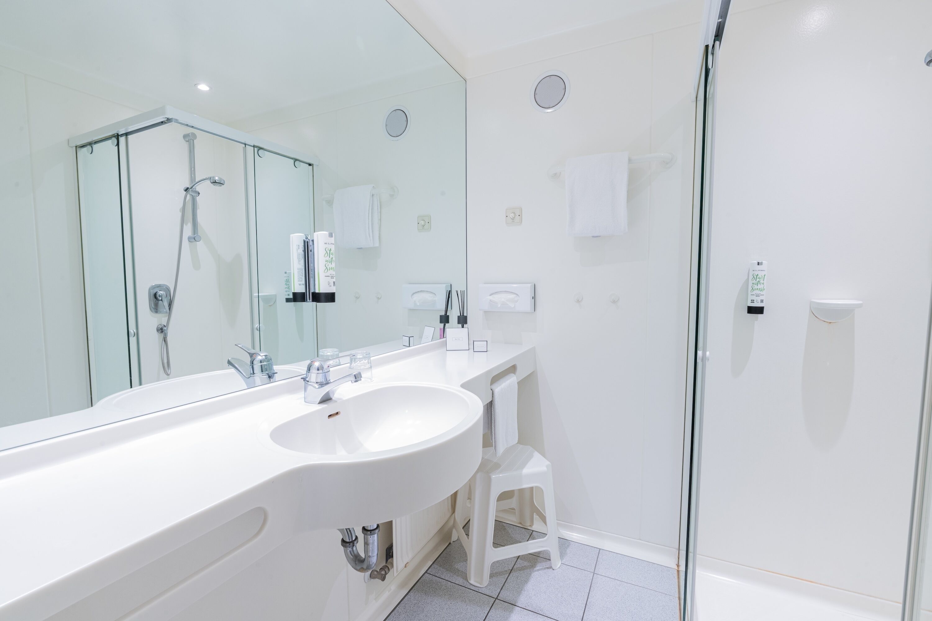 A modern bathroom with a large mirror, a white sink, a glass shower enclosure, and a white towel on the wall.