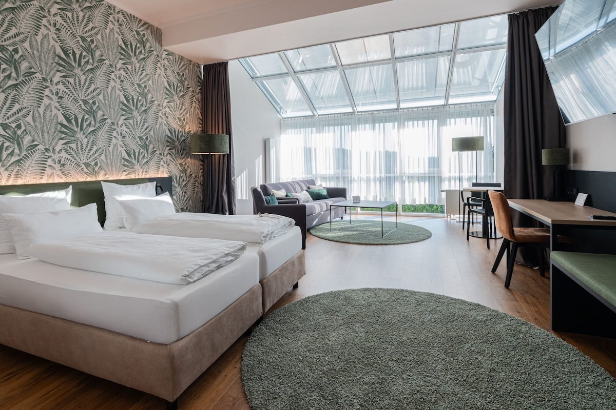 A hotel room with a large bed, a desk, a sofa, and a view of the city through large windows.