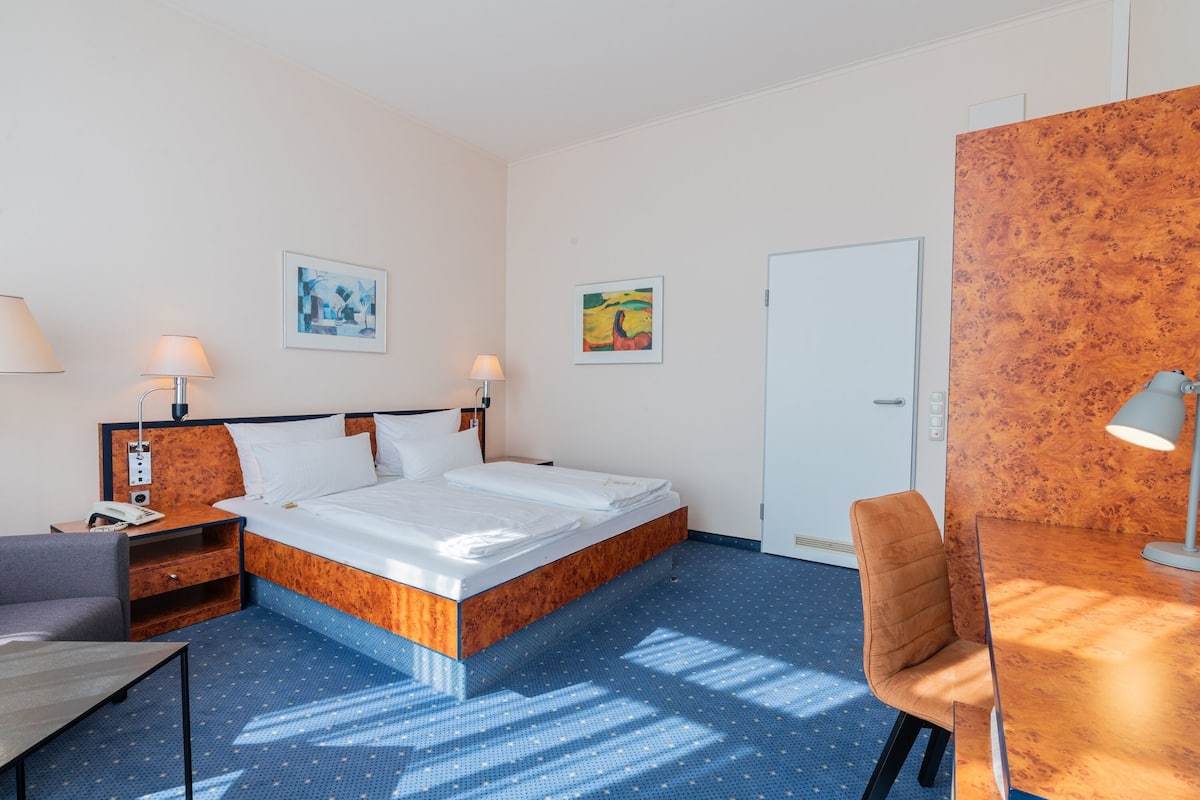 A hotel room with a bed, bedside tables, a desk, a chair, and artwork on the walls.