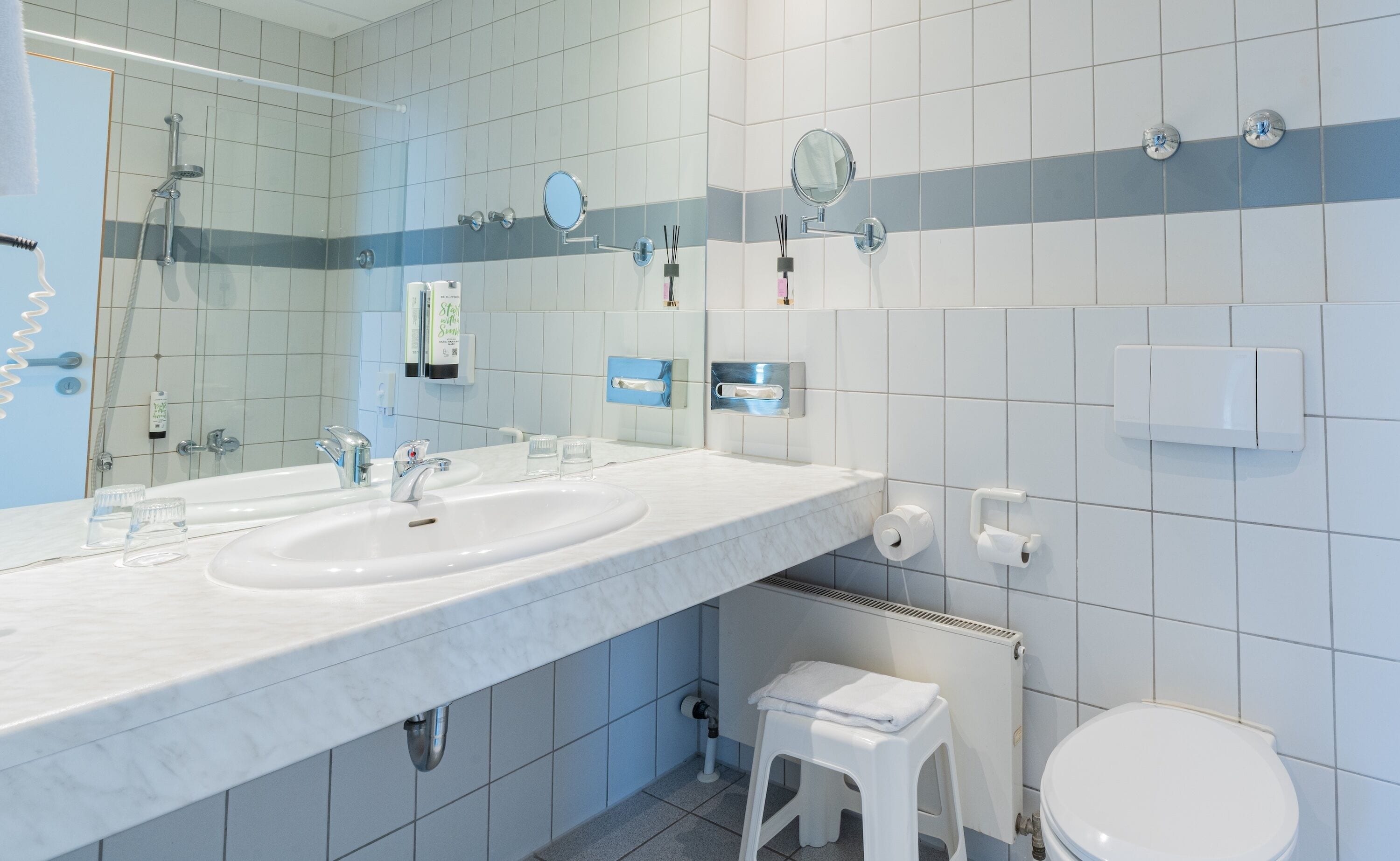 Een moderne badkamer met een dubbele wastafel, toilet, een klein krukje en een handdoekrek.