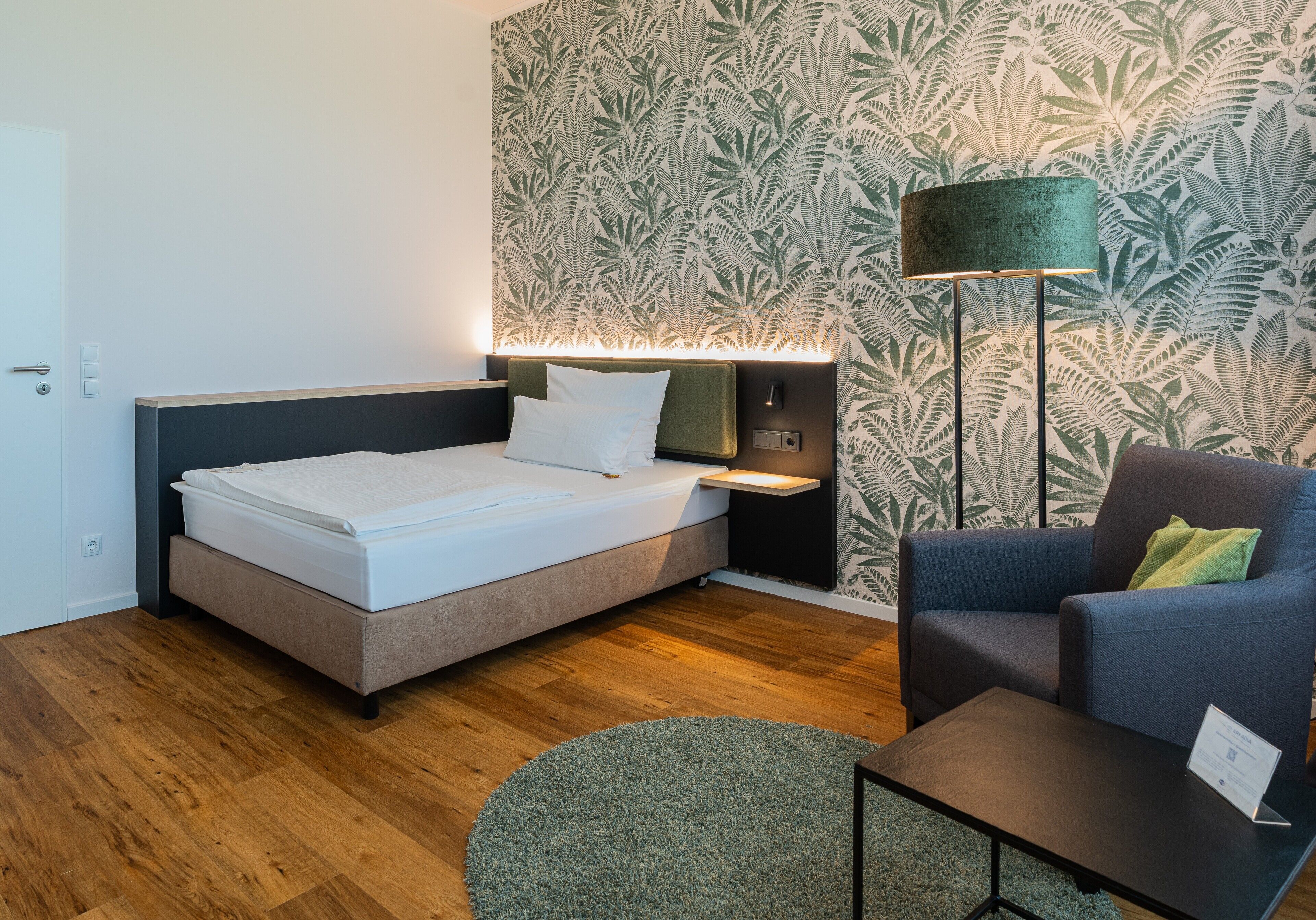 A modern hotel room with a bed, a nightstand, a lamp, a chair, and a small table.