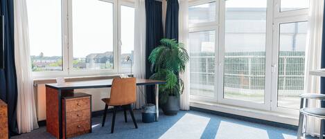 A room with a desk, chair, potted plant, and large windows.