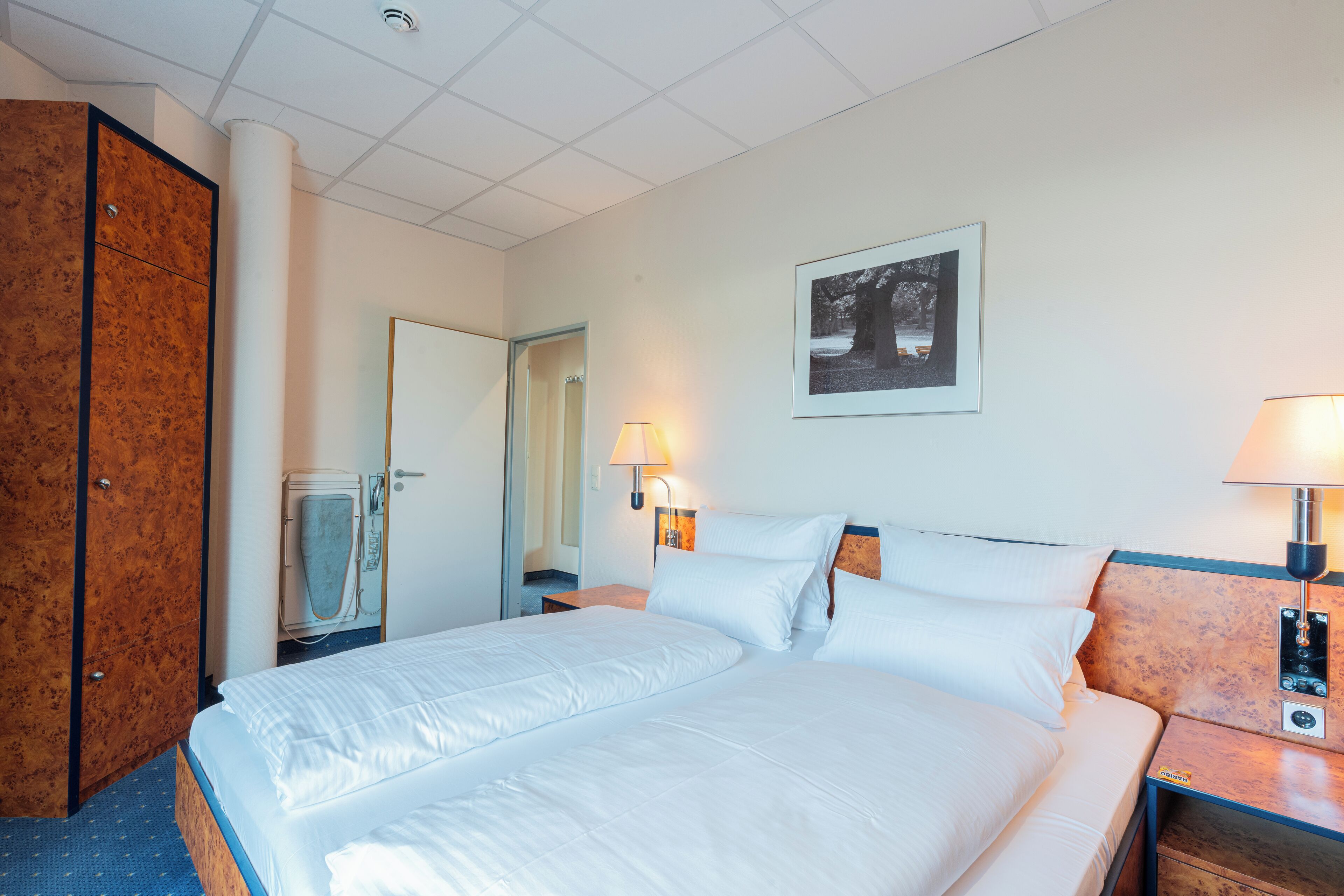 Une chambre d’hôtel avec un grand lit, une armoire en bois, une salle de bain avec une serviette suspendue, un tableau encadré au mur et une table de chevet avec une lampe.