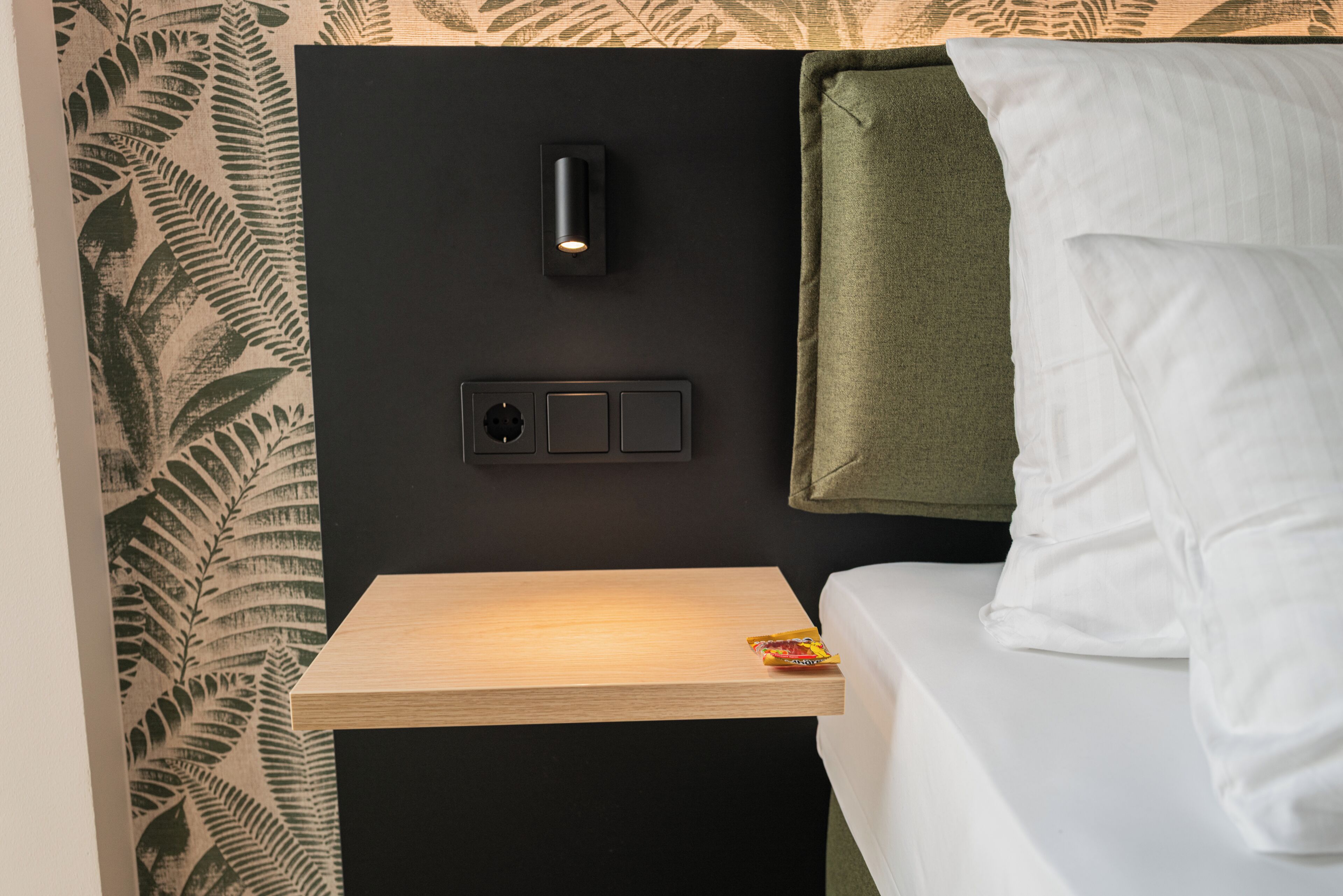 A bed with white sheets, a green towel, a nightstand with a light switch, and a wall with a fern pattern.