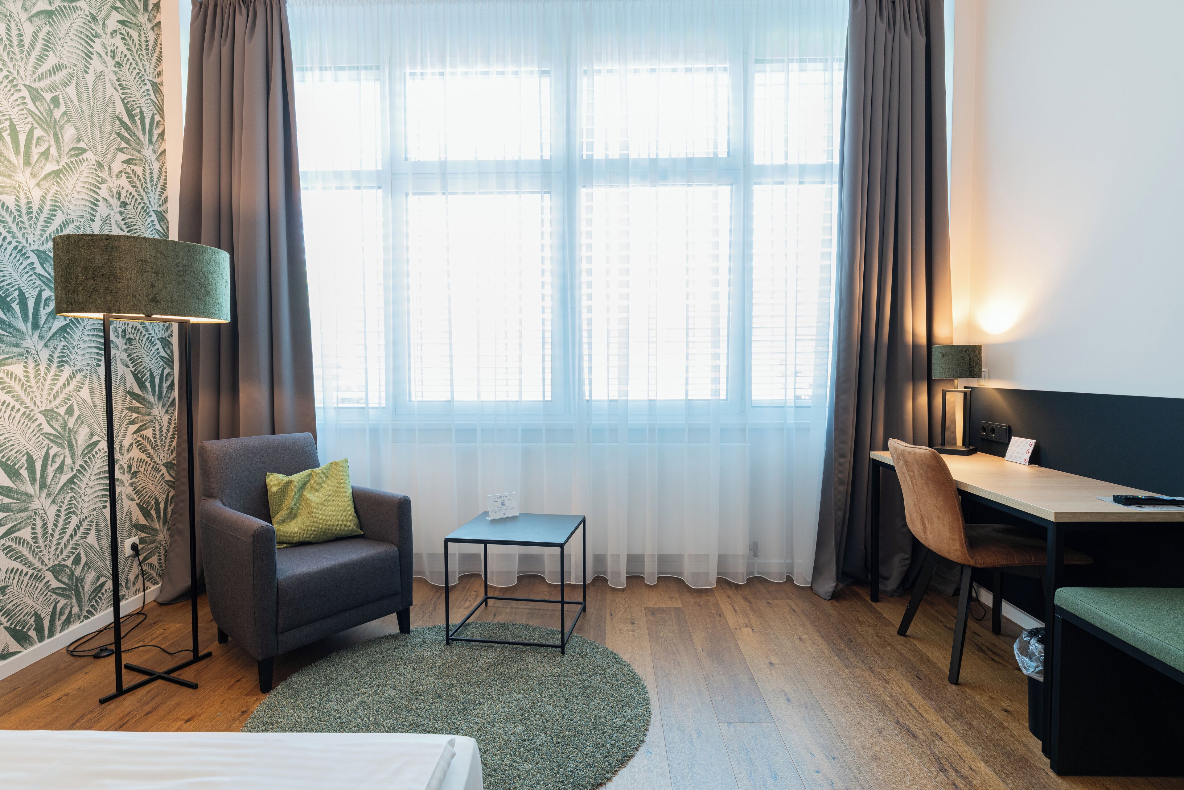 A hotel room with a bed, a desk, a chair, a small table, a floor lamp, and a window with curtains.