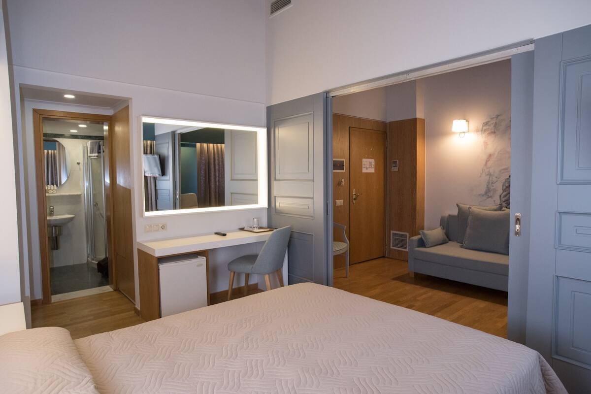 A hotel room with a bed, a desk with a chair, a sofa, a mirror, and a shower area.
