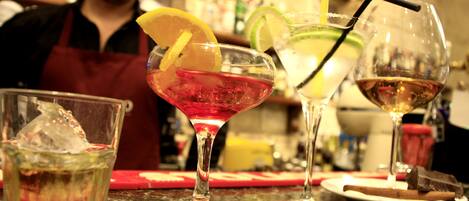 Un bar con tres bebidas, una persona con delantal y varias botellas al fondo.
