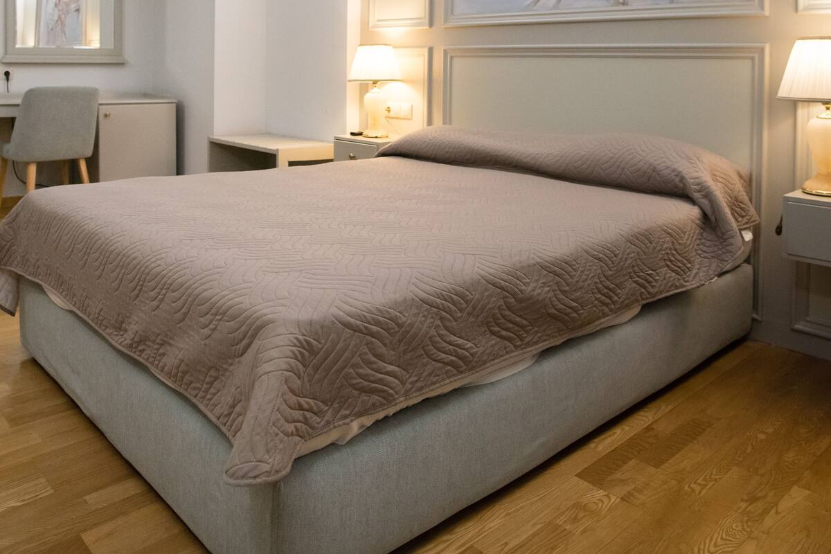A neatly arranged bedroom with a large bed, a desk, and a chair. There are two bedside lamps and a painting on the wall.
