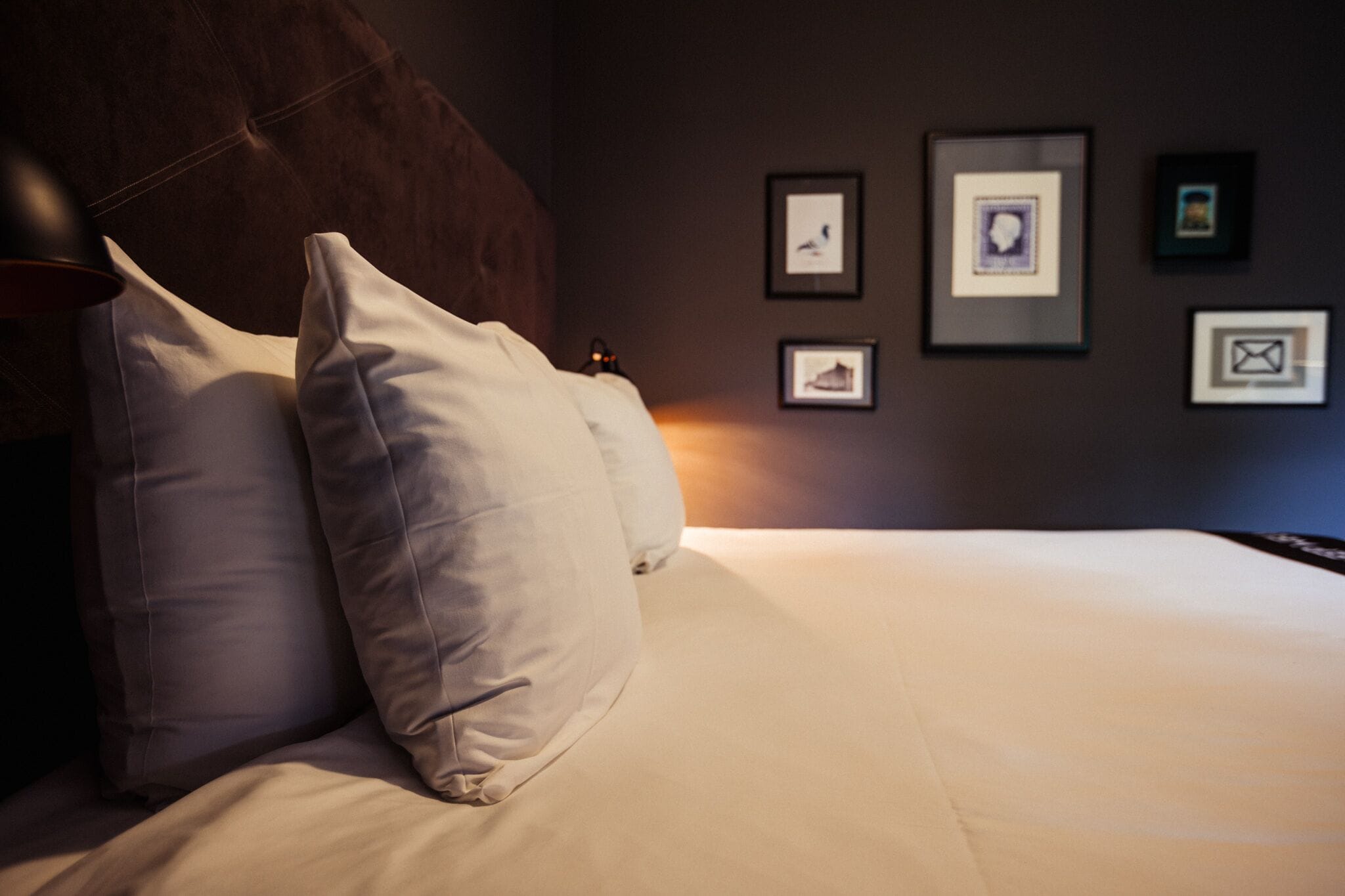 Een netjes opgemaakt bed met witte lakens en twee kussens, een donker hoofdbord en een muur met ingelijste foto's.