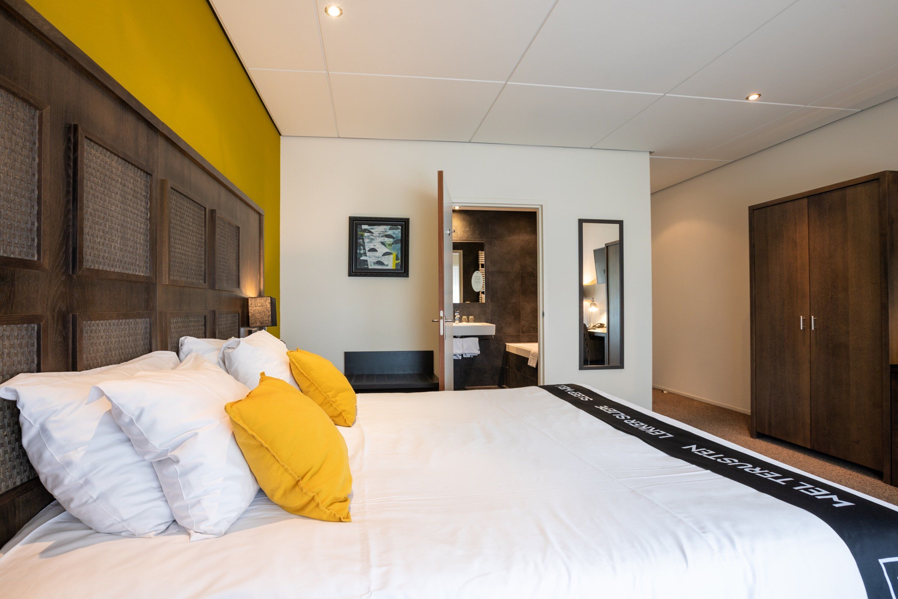 Een moderne hotelkamer met een groot bed, een houten hoofdeinde, een gele accentmuur en uitzicht op de badkamer.