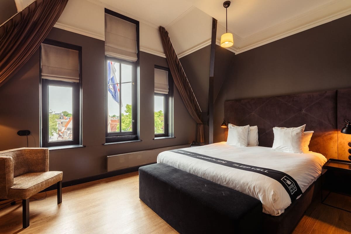 Ein modernes Schlafzimmer mit einem großen Bett, einem Sessel und Blick auf die Stadt durch große Fenster.