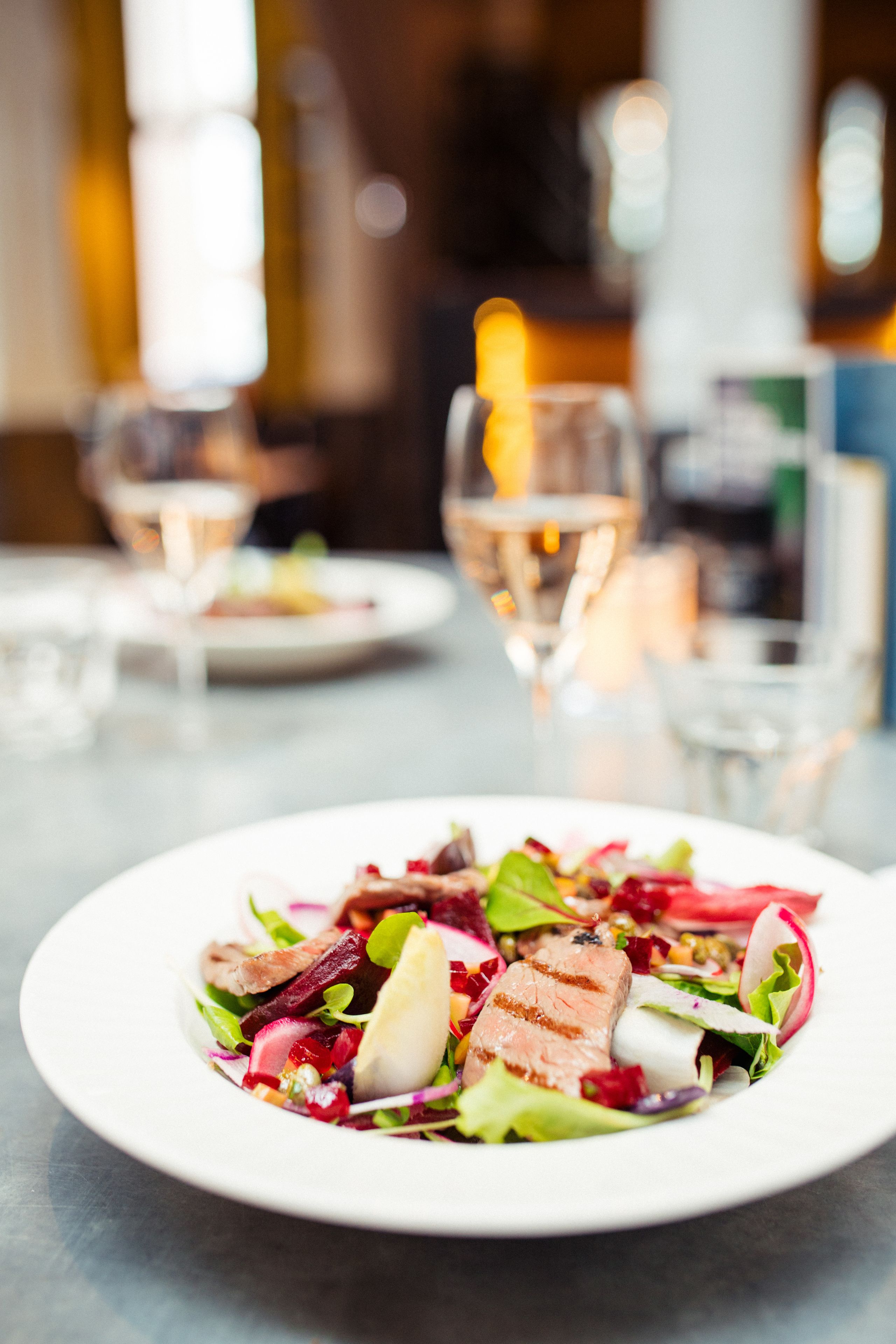 A plate of food with various vegetables and meat, set on a table with glasses of wine in the background.