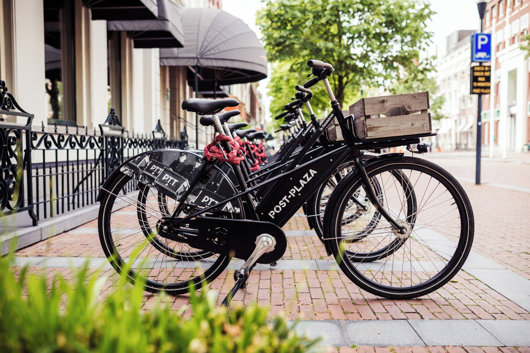 A black Post-PLAZA bicycle parked on a brick sidewalk with a wooden crate on the back.