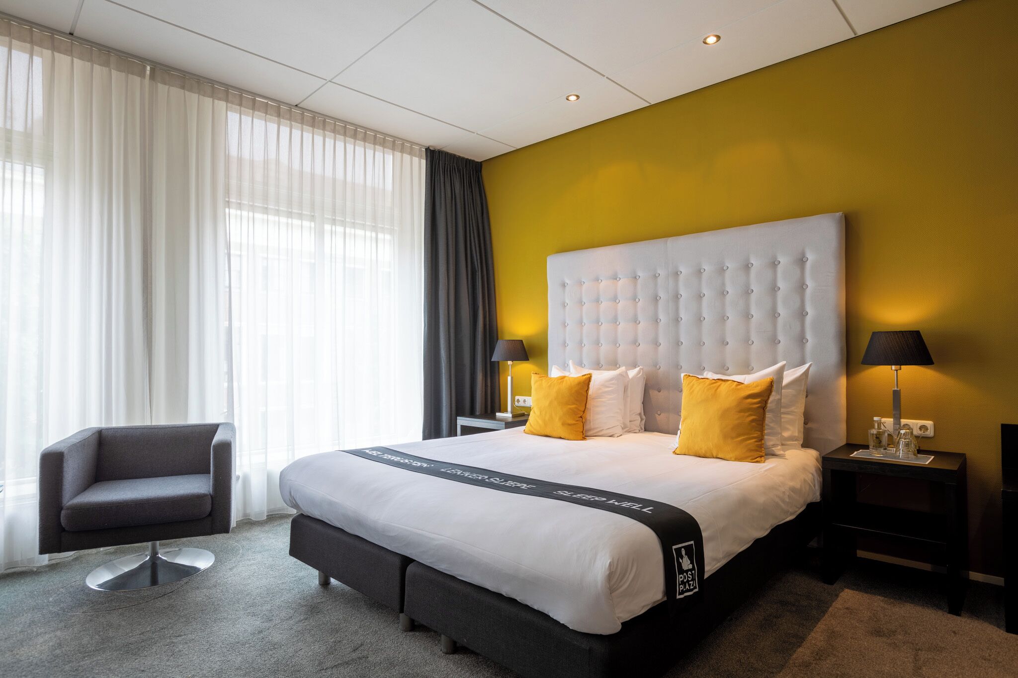 Ett modernt hotellrum med en stor säng, en grå fåtölj och ett nattduksbord med lampor.