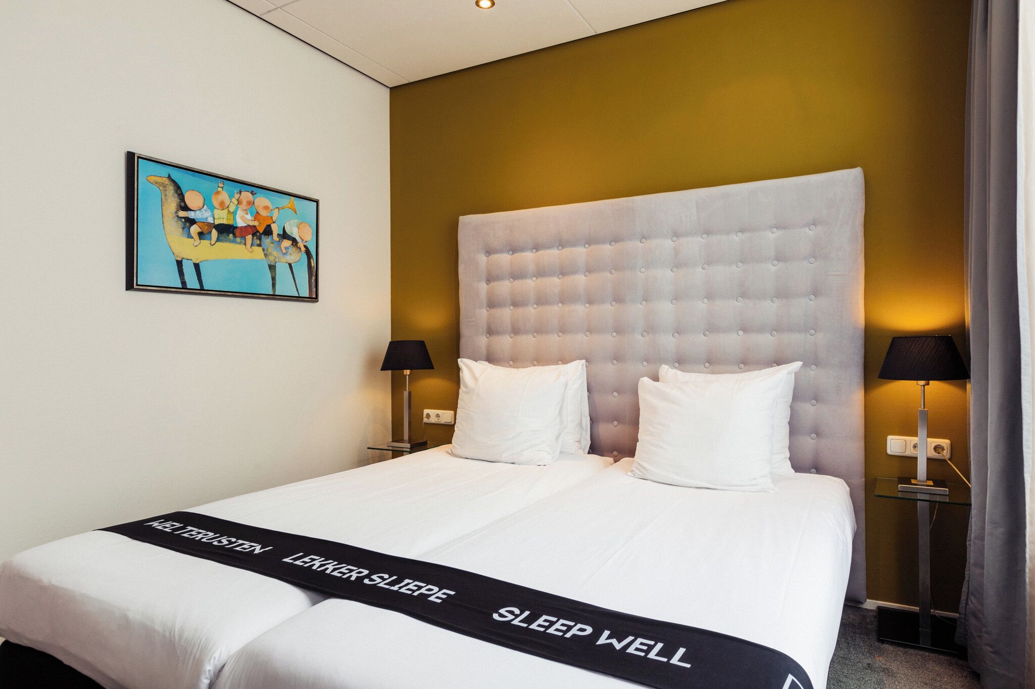 Una habitación de hotel con una cama grande, dos lámparas de noche, un cuadro en la pared y una colcha con texto.