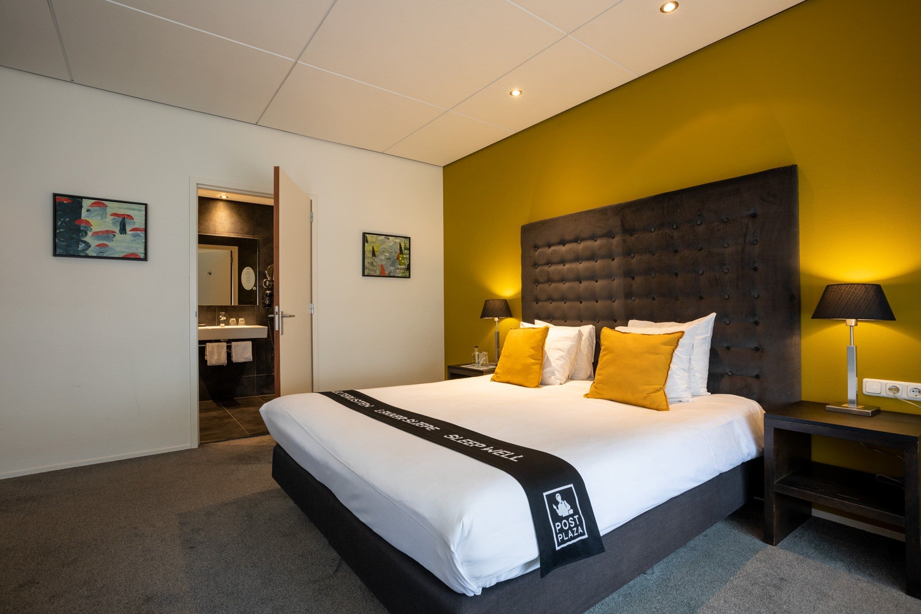 Een hotelkamer met een groot bed, een nachtkastje met een lamp en een badkamer zichtbaar door een open deur.