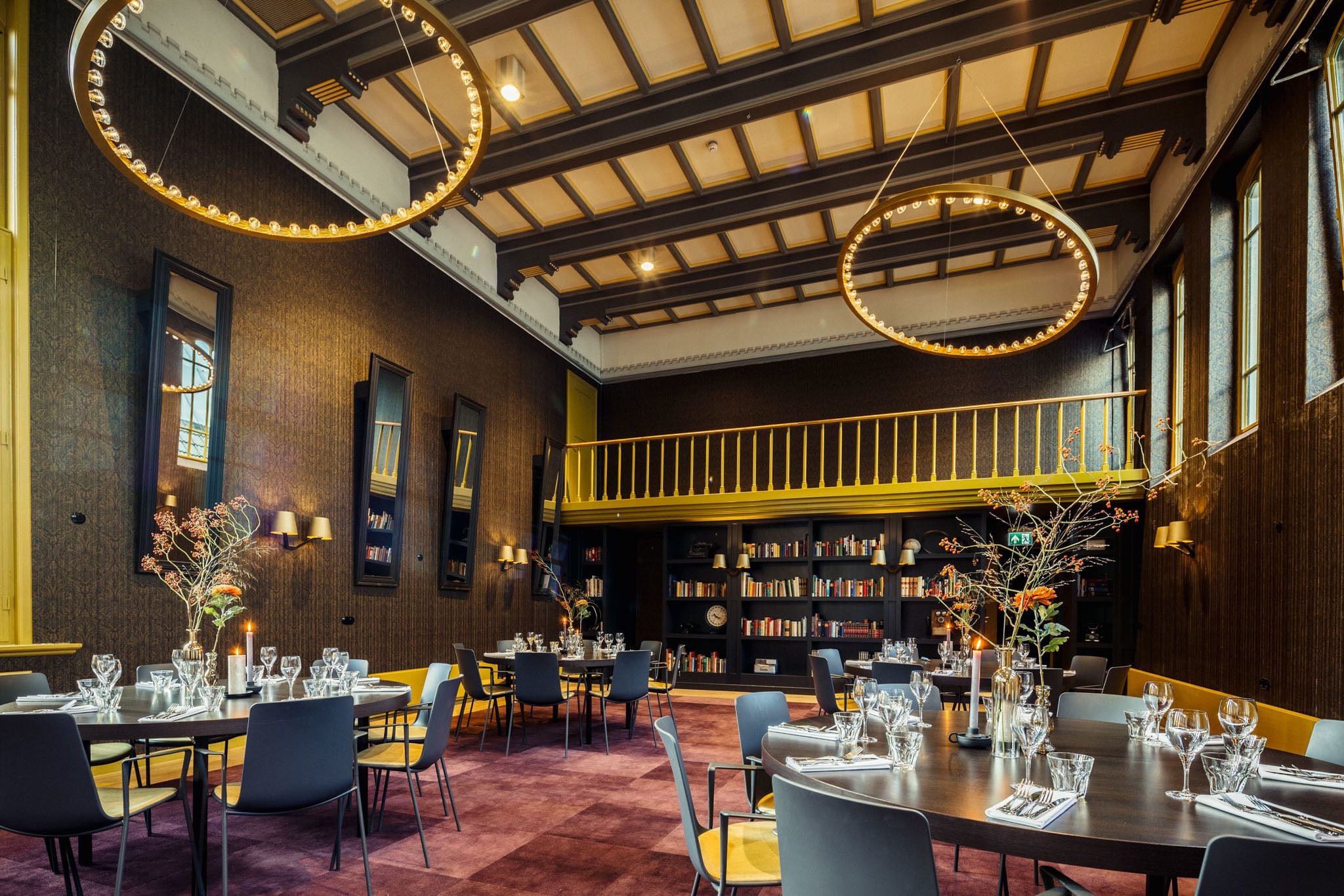 A dining area with tables set for service, a bookshelf, and a staircase leading to an upper level.