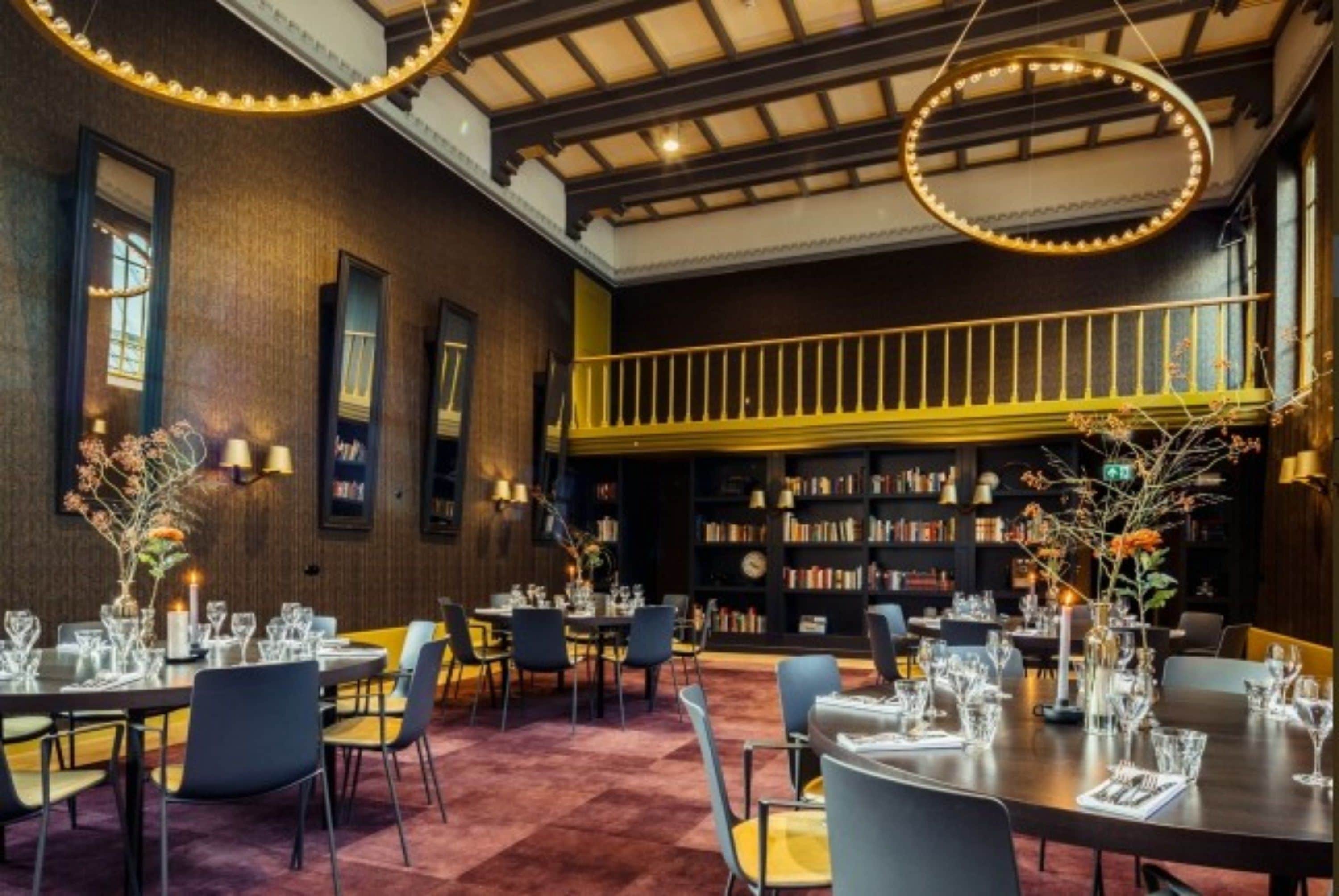 A dining area with tables set for a meal, a bookshelf in the background, and a balcony with a railing.